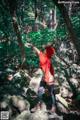 A woman in a red hoodie standing on a rock in the woods.