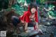 A woman in a red hooded outfit crouching on a rock in the woods.