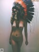 A naked woman wearing an indian headdress standing in front of a wall.