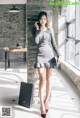 A woman in a gray dress standing next to a brick wall.