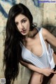 A woman with long black hair posing in front of a wall.