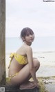 A woman in a yellow bikini sitting on a rock by the ocean.